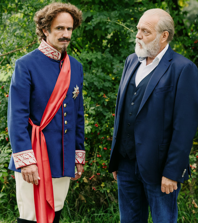 "Der Alte - Der Mondkönig": Robert Obermüller (Felix Hellmann), in der Rolle von Ludwig II. eingekleidet, steht mit Richard Voss (Jan-Gregor Kremp) auf einer Wiese im Park. Die beiden sprechen miteinander. Robert Obermüller dreht sich ertappt weg von Voss.