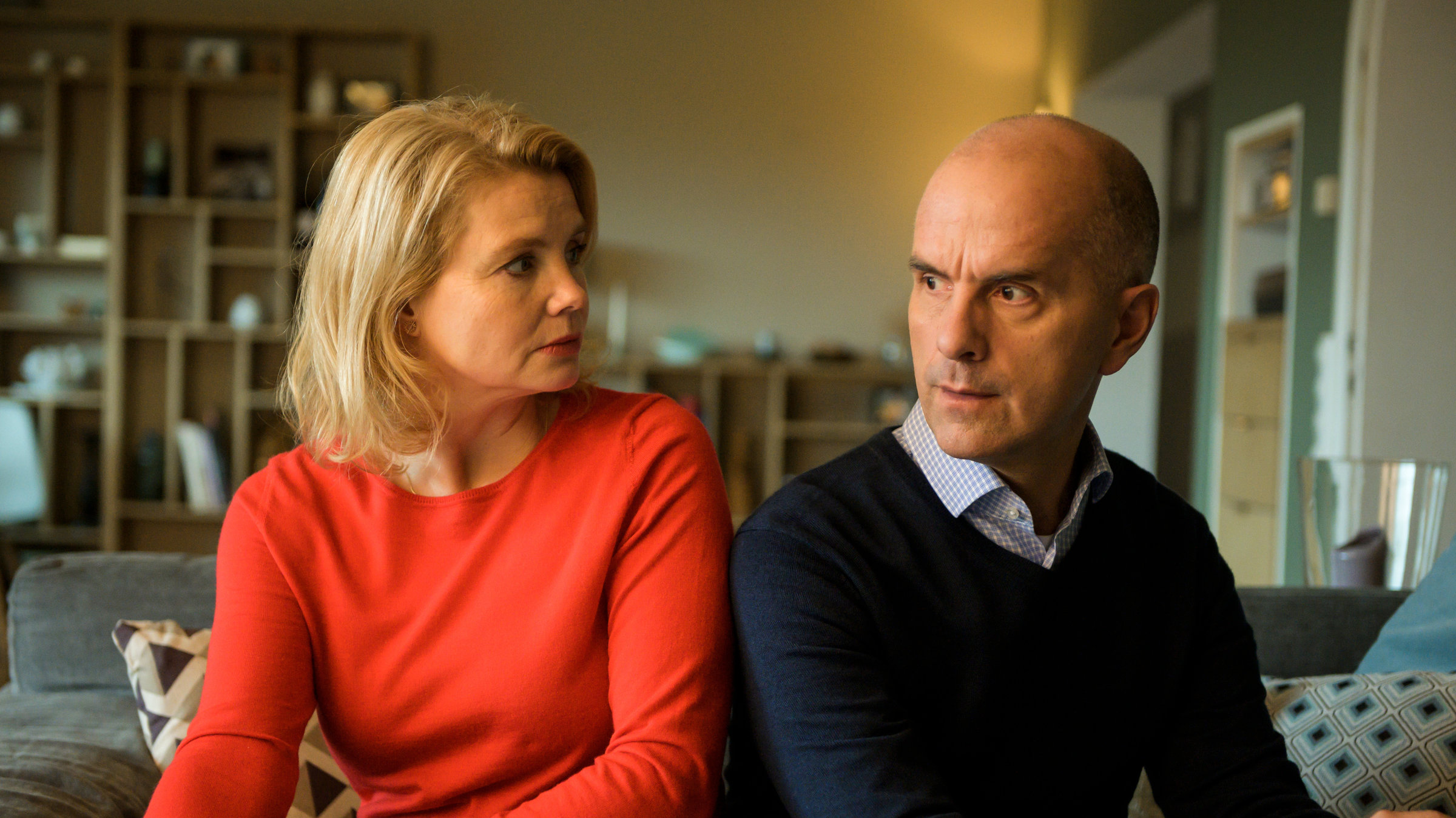 "Merz gegen Merz - Auszeiten": Anne (Annette Frier) sitzt in einem roten Pulli neben ihrem Mann Erik (Christoph Maria Herbst). Sie sitzen nebeneinander in ihrem Wohnzimmer auf der Couch. Beide schauen irritiert.