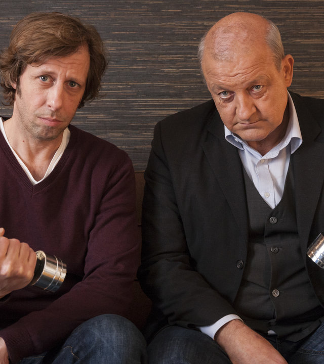 "Wilsberg - Bauch, Beine, Po": Ekki Talkötter (Oliver Korittke) und Georg Wilsberg (Leonard Lansink) sitzen nebeneinander auf einem Sofa. Beide haben jeweils eine Hantel in der Hand.