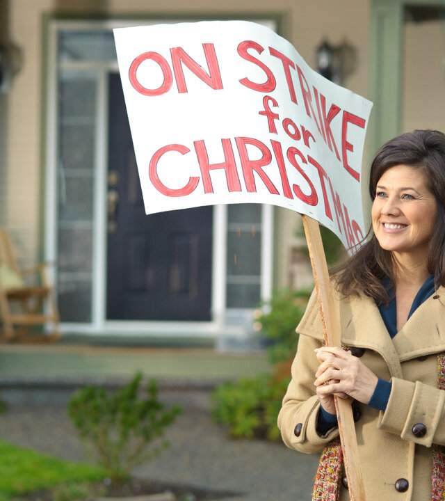"Der Weihnachtsstreik": Joy Robertson (Daphne Zuniga) steht im Mantel vor ihrem Haus. Sie lächelt und trägt eine Schild in den Händen, auf dem "On strike for christmas" steht.