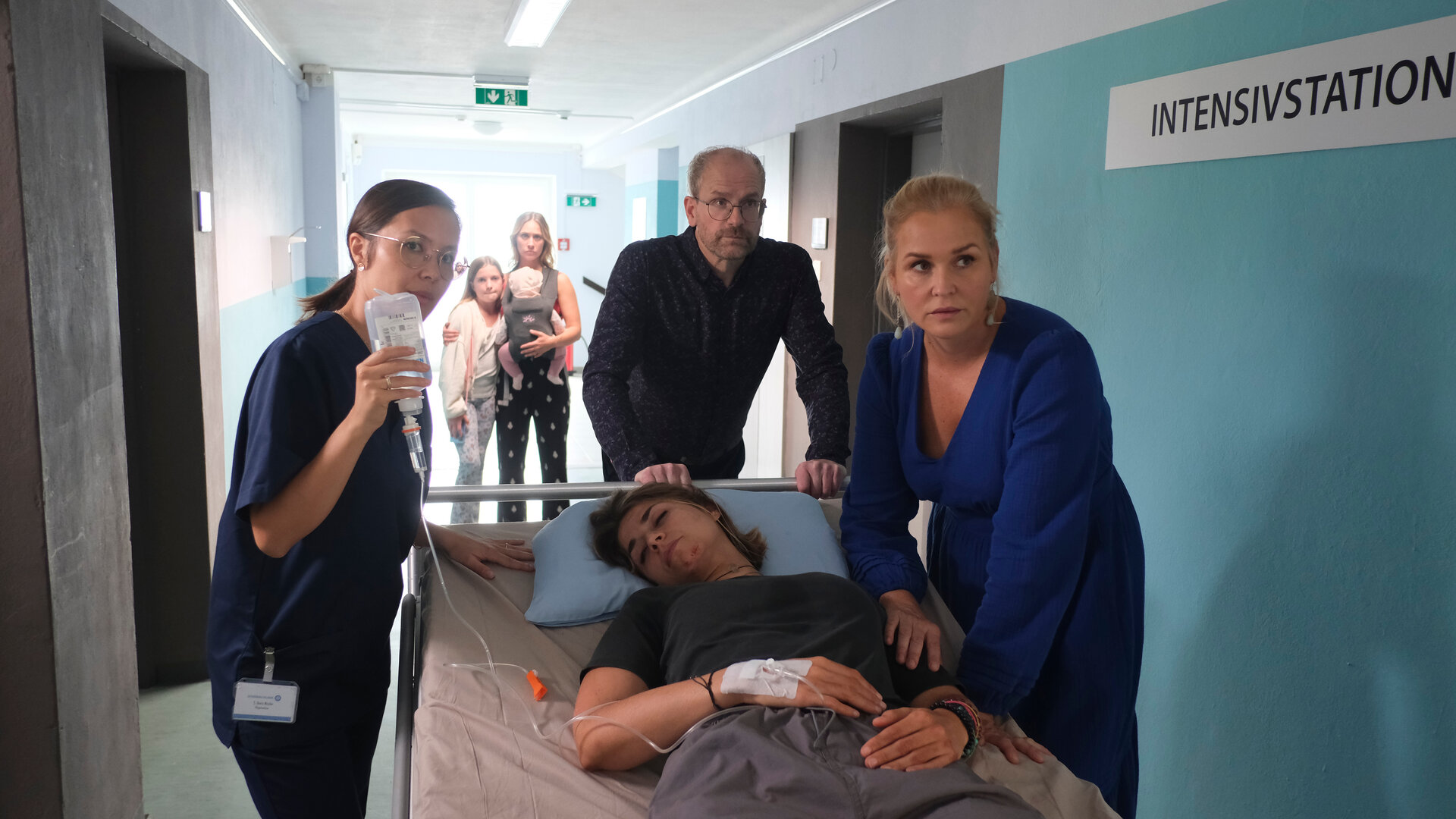 "Lena Lorenz - Blinder Passagier": Dr. Stefan Keller (Thomas Limpinsel) und Lena Lorenz (Judith Hoersch) schieben ein Krankenbett, auf dem Enissa Heydari (Jaëla Probst) liegt, durch einen Krankenhausflur zur Intensivstation. Eine Pflegerin (Komparsin) geht nebenher und hält eine Infusionsflasche, deren Nadel an Enissas Handrücken angebracht ist, in die Höhe. Im Hintergrund steht Mina Heydari (Sina Tkotsch) mit Baby Ella und Tochter Lara (Martha Felipa Abel).