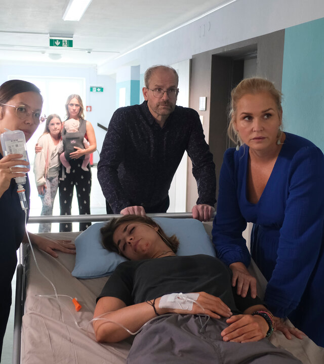 "Lena Lorenz - Blinder Passagier": Dr. Stefan Keller (Thomas Limpinsel) und Lena Lorenz (Judith Hoersch) schieben ein Krankenbett, auf dem Enissa Heydari (Jaëla Probst) liegt, durch einen Krankenhausflur zur Intensivstation. Eine Pflegerin (Komparsin) geht nebenher und hält eine Infusionsflasche, deren Nadel an Enissas Handrücken angebracht ist, in die Höhe. Im Hintergrund steht Mina Heydari (Sina Tkotsch) mit Baby Ella und Tochter Lara (Martha Felipa Abel).