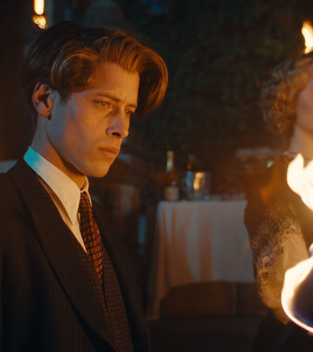 "This Is Not a Murder Mystery - Das Hummer-Telefon": René Magritte (Pierre Gervais) und Lee Miller (Florence Hall) stehen nebeneinander im Dunkeln. Im Vordergrund brennt eine Fackel, hinter den beiden sind weitere Lichter erkennbar.