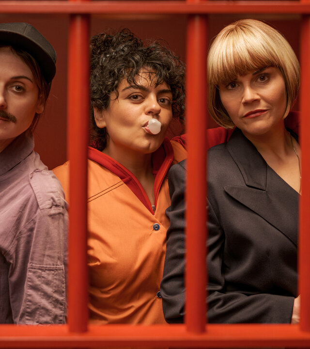 "Gar kein Geld macht auch nicht glücklich": Lesley (Julia Becker), Olivia (Sara Fazilat) und Kim (Katharina Wackernagel) stehen lässig nebeneinander in einem Raum mit knallroten Wänden. Vor ihnen sind rote Gitterstäbe, die die drei Frauen darin einsperren. Olivia formt mit dem Mund eine Kaugummiblase und legt ihren Arm um Kims Schultern. Diese hält eine Pistole in der Hand.