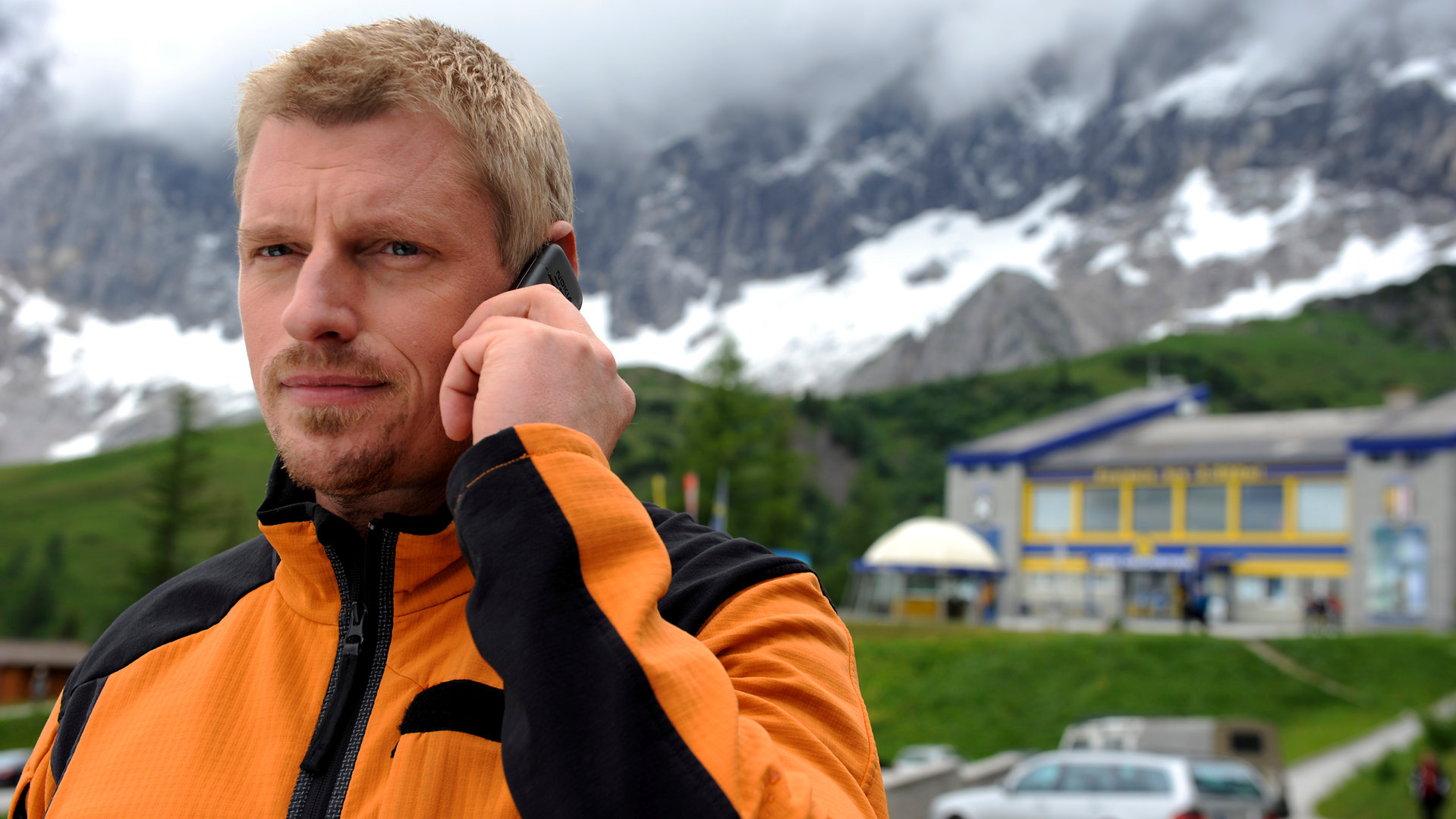 "Die Bergretter: Steinschlag (1)": Andreas (Martin Gruber) steht mit einem Telefon am linken Ohr vor einer Berg-Kulisse.