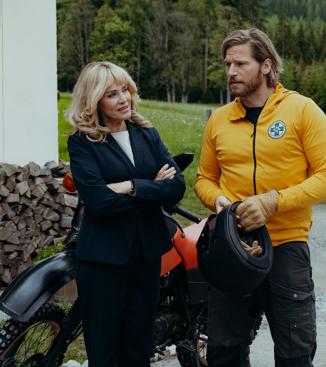 "Die Bergretter: Stille Wasser": Auf Emilies Hof: Markus Kofler (Sebastian Ströbel) steht neben einem Motorrad, hält einen Helm in den Händen und blickt fragend zu Elisa Rödder (Sonja Kirchberger). Diese steht, die Arme vor der Brust verschränkt, neben ihm und wirkt triumphierend.