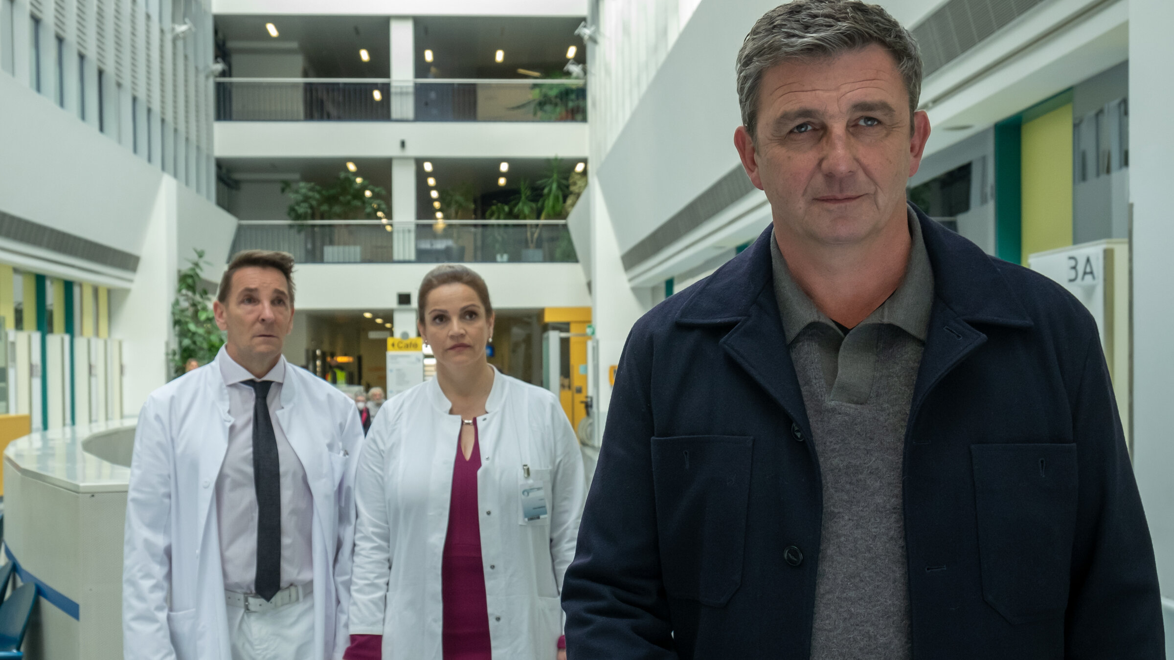 "Der Bergdoktor - Der Weg zurück (2)": Im Krankenhaus: Hand in Hand stehen Dr. Alexander Kahnweiler (Mark Keller) und Dr. Vera Fendrich (Rebecca Immanuel) zusammen. Sie blicken traurig Dr. Martin Gruber (Hans Sigl) nach, der sich mit einem Lächeln auf dem Gesicht von beiden entfernt.