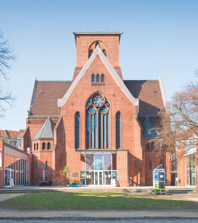"Evangelischer Gottesdienst - Ein Platz bei Gott": Außenansicht der aus rötlichem Backstein erbauten Genezareth-Kirche in Berlin-Neukölln. Vor der Kirche befindet sich ein weitläufiger Platz mit einer Rasenfläche und Bäumen.
