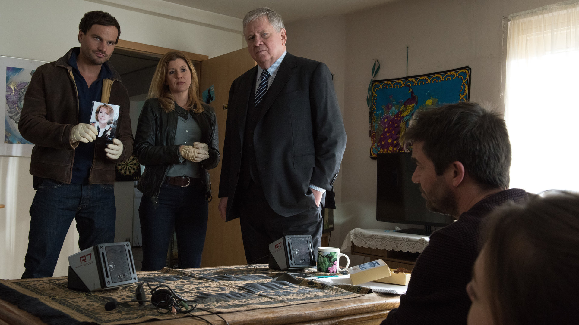 "Der Staatsanwalt - Die Enkelin": Christian Schubert (Simon Eckert), Kerstin Klar (Fiona Coors) und Bernd Reuther (Rainer Hunold) stehen in einem Zimmer vor Peter Blass (Thomas Scharff) und Paula Blass (Michelle Barthel). Die beiden sitzen hintereinander an einem Tisch und schauen die drei vor ihnen Stehenden an. Schubert zeigt ihnen das Foto einer Frau.