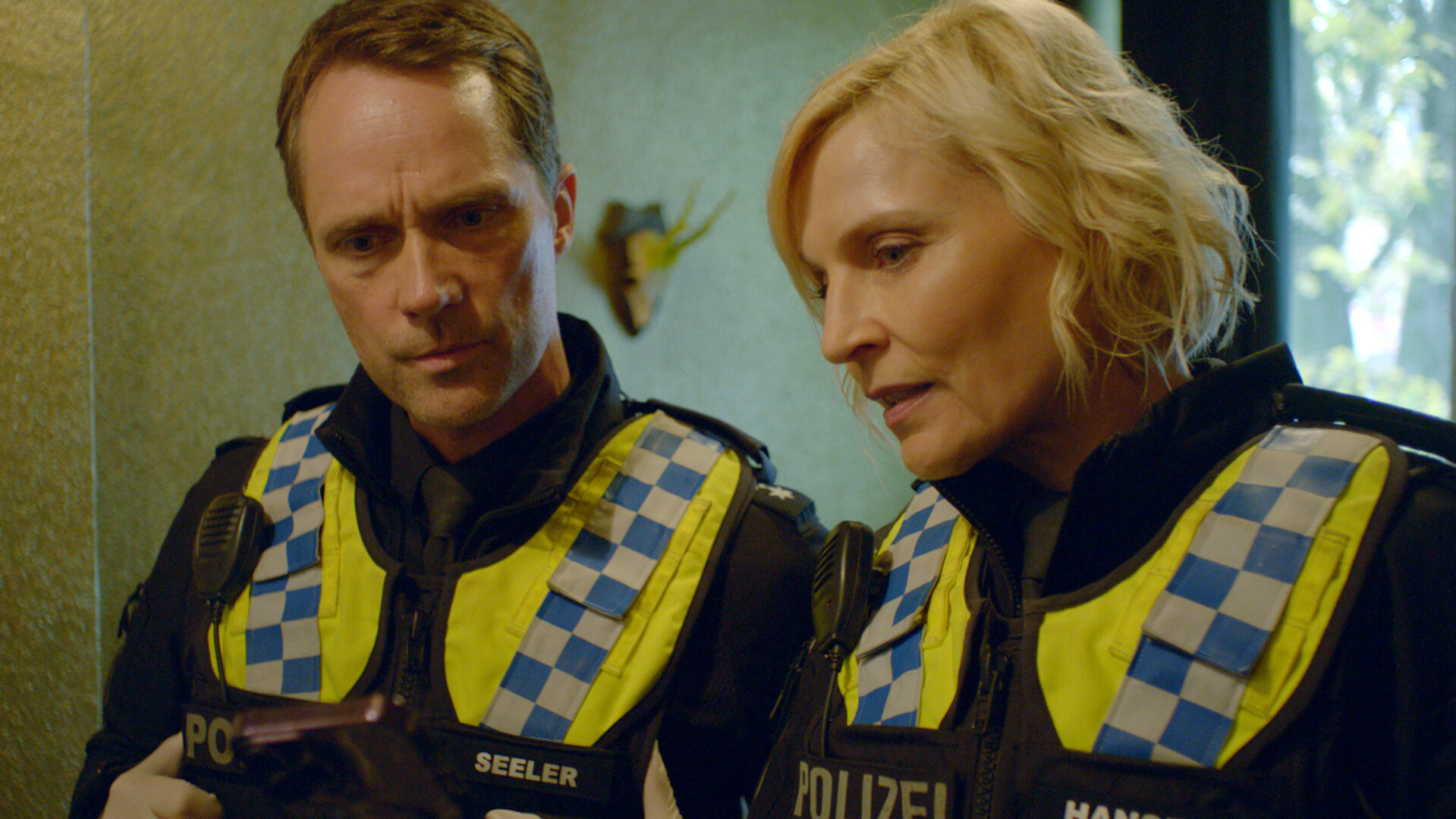 "Notruf Hafenkante - Toxisch": Großaufnahme: Polizistin Melanie Hansen (Sanna Englund) und ihr Kollege Mattes Seeler (Matthias Schloo) stehen dicht nebeneinander in einem Raum. Beide tragen Latexhandschuhe. Melanie hält ein Handy in den Händen auf das beide schauen.