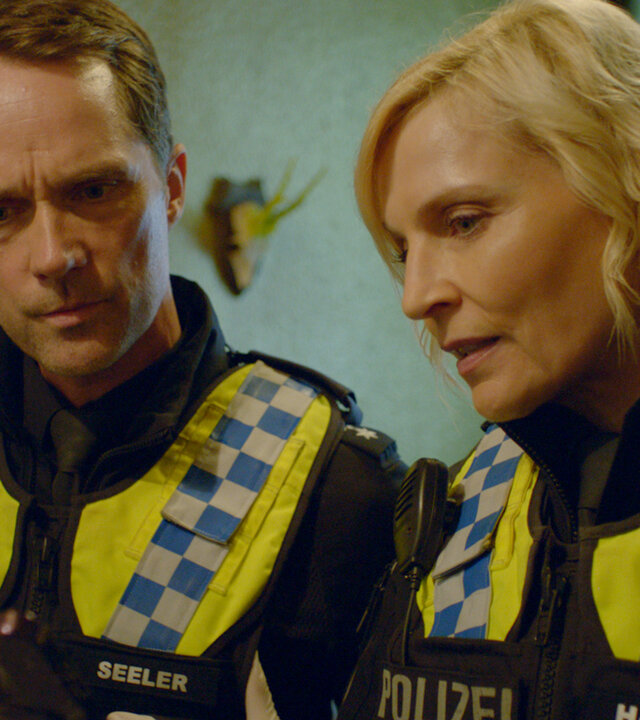 "Notruf Hafenkante - Toxisch": Großaufnahme: Polizistin Melanie Hansen (Sanna Englund) und ihr Kollege Mattes Seeler (Matthias Schloo) stehen dicht nebeneinander in einem Raum. Beide tragen Latexhandschuhe. Melanie hält ein Handy in den Händen auf das beide schauen.