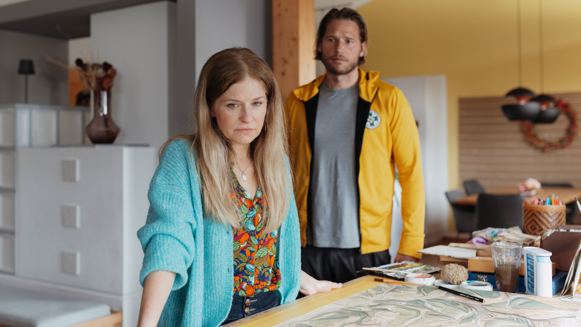 "Die Bergretter: Stimmen der Vergangenheit": Im Haus der Familie HÃ¶ller: Mit traurigem Blick stÃ¼tzt sich Isabel HÃ¶ller (Birte Hanusrichter) an einem Arbeitstisch ab. Auf der dort ausgebreiteten groÃformatige Zeichnung einer geografischen Karte liegt ein Handy. Im Hintergrund steht Bergretter Markus Kofler (Sebastian StrÃ¶bel) und blickt mitfÃ¼hlend zu Isabel.