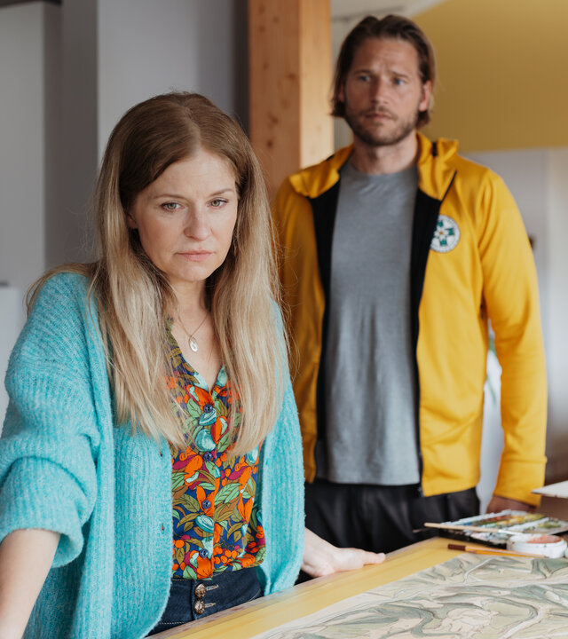"Die Bergretter: Stimmen der Vergangenheit": Im Haus der Familie HÃ¶ller: Mit traurigem Blick stÃ¼tzt sich Isabel HÃ¶ller (Birte Hanusrichter) an einem Arbeitstisch ab. Auf der dort ausgebreiteten groÃformatige Zeichnung einer geografischen Karte liegt ein Handy. Im Hintergrund steht Bergretter Markus Kofler (Sebastian StrÃ¶bel) und blickt mitfÃ¼hlend zu Isabel.