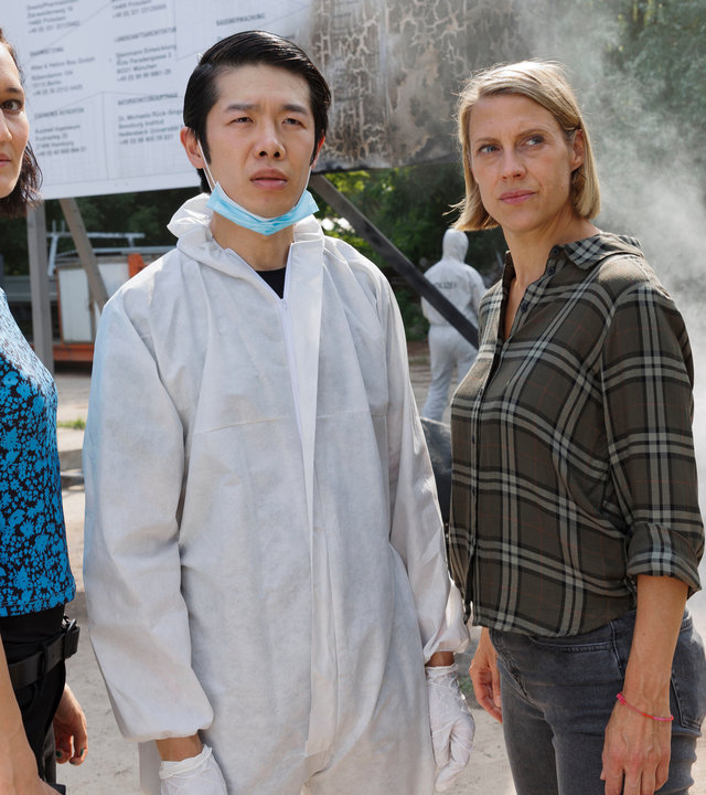 "SOKO Potsdam - Tatütata": Auf einem Firmengelände stehen Pauline Hobrecht (Agnes Decker), Spurensicherer Thomas Brandner (Yung Ngo) und Tamara Meurer (Anja Pahl). Im Hintergrund kümmern sich Spurensicherer und Feuerwehr um die durch den Brandanschlag verursachten Schäden.