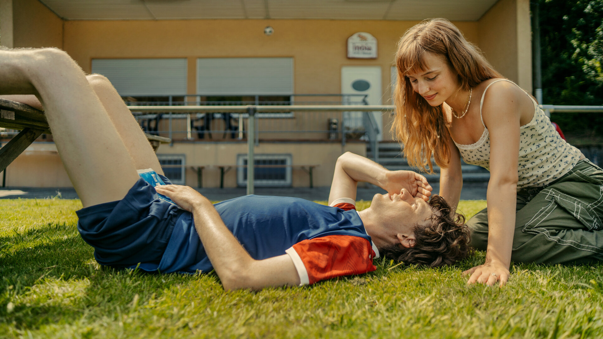 "Tschappel - Papa (T)ante Sportplatz": Carlo Brenner (Jeremias Meyer) liegt auf dem Sportplatz auf dem Rasen, während Pia Schneider (Mina-Giselle Rüffer) sich über ihn lehnt.
