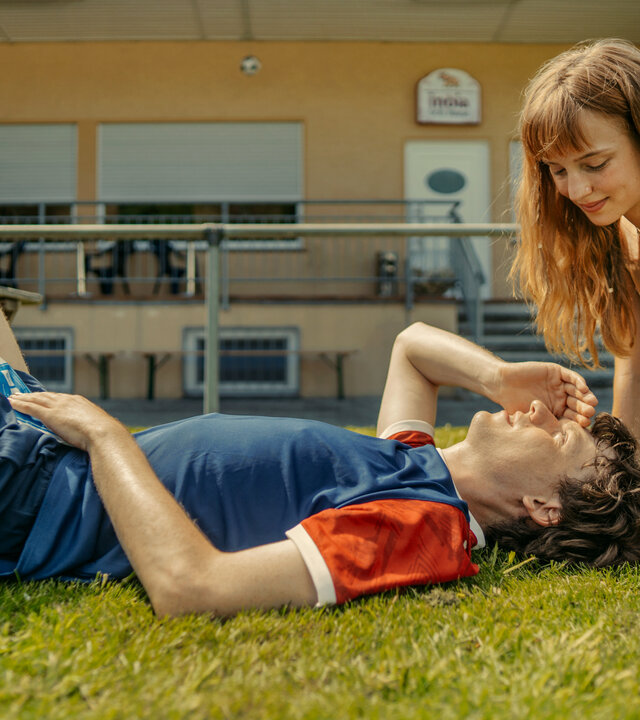 "Tschappel - Papa (T)ante Sportplatz": Carlo Brenner (Jeremias Meyer) liegt auf dem Sportplatz auf dem Rasen, während Pia Schneider (Mina-Giselle Rüffer) sich über ihn lehnt.