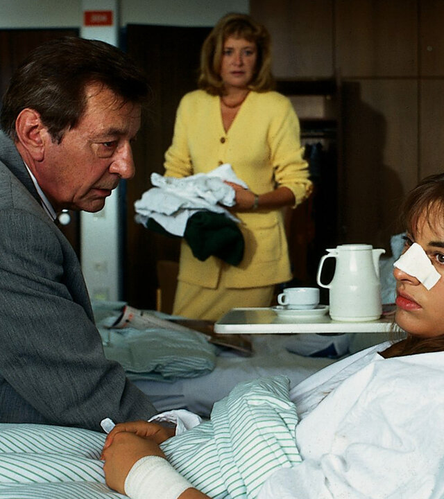 "Alle meine Töchter - Tante Mathilde greift ein": Sylvie von Wettenberg (Julia Dahmen) liegt abwesend in ihrem Krankenhausbett. Neben ihr sitzt ihr Vater Dr. Berthold Sanwaldt (Günter Mack), der besorgt auf sie schaut. Margot Sanwaldt (Jutta Speidel) steht in der Mitte des Raumes und hält Kleidung in ihren Händen.
