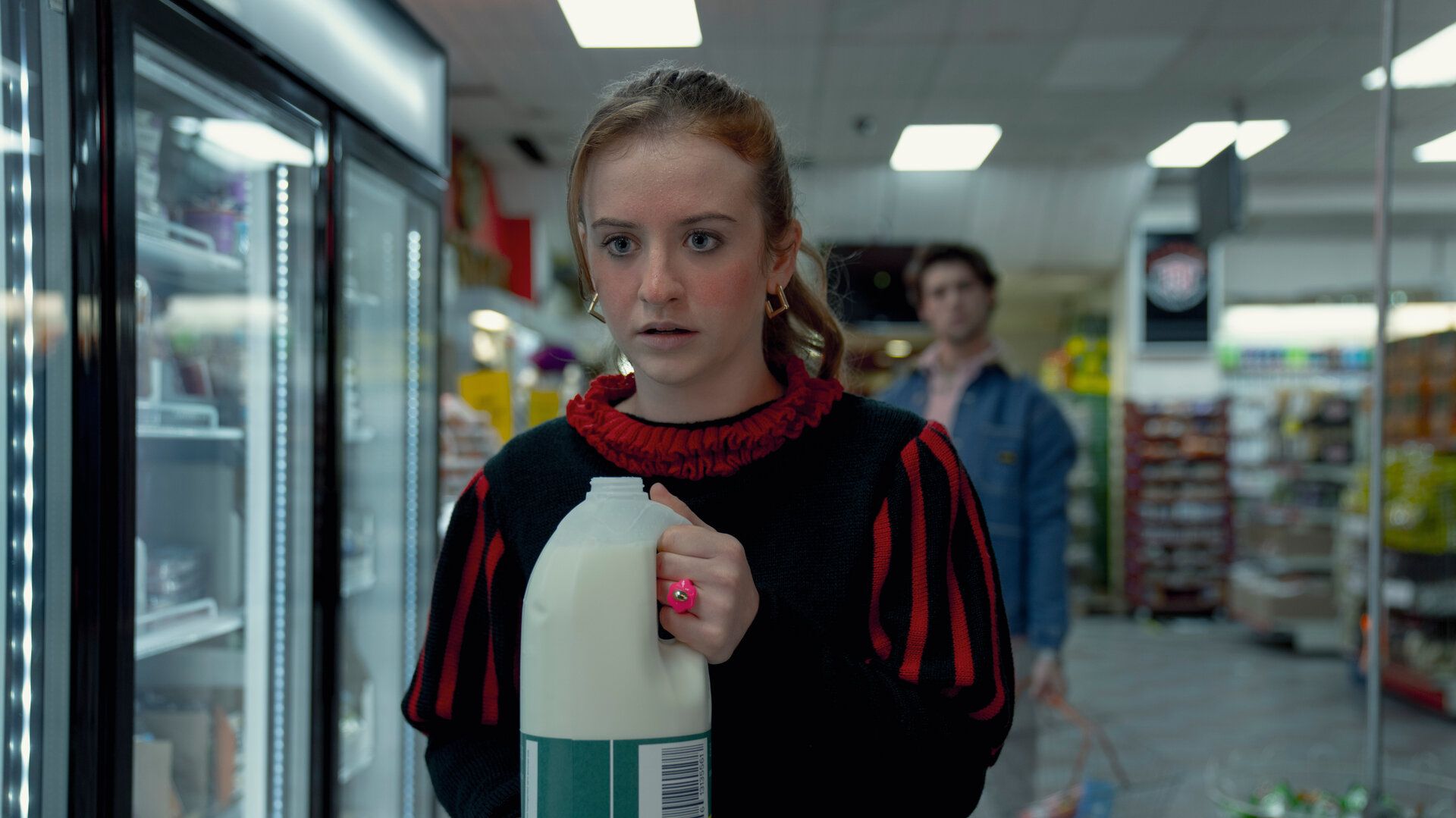 "Extraordinary - Wundermittel": Jen (Máiréad Tyers) steht mit einer geöffneten Milchflasche in den Händen in einem Supermarkt, während ihr Blick angespannt und leicht erschrocken ist. Hinter ihr steht Luke (Ned Porteous), der sie aus einiger Entfernung beobachtet und einen Einkaufskorb in der Hand hält.