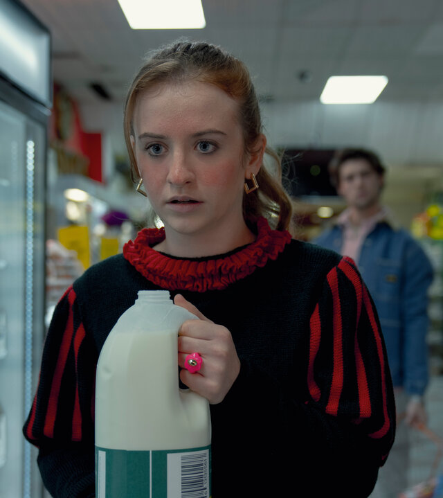 "Extraordinary - Wundermittel": Jen (Máiréad Tyers) steht mit einer geöffneten Milchflasche in den Händen in einem Supermarkt, während ihr Blick angespannt und leicht erschrocken ist. Hinter ihr steht Luke (Ned Porteous), der sie aus einiger Entfernung beobachtet und einen Einkaufskorb in der Hand hält.