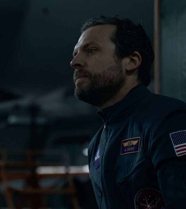 "Infiniti - Turm des Schweigens": Anthony Kurz (Lex Shrapnel) steht in einem dunklen Hangar und schaut nachdenklich in die Ferne. Dabei trägt er seinen NASA-Jumpsuit.