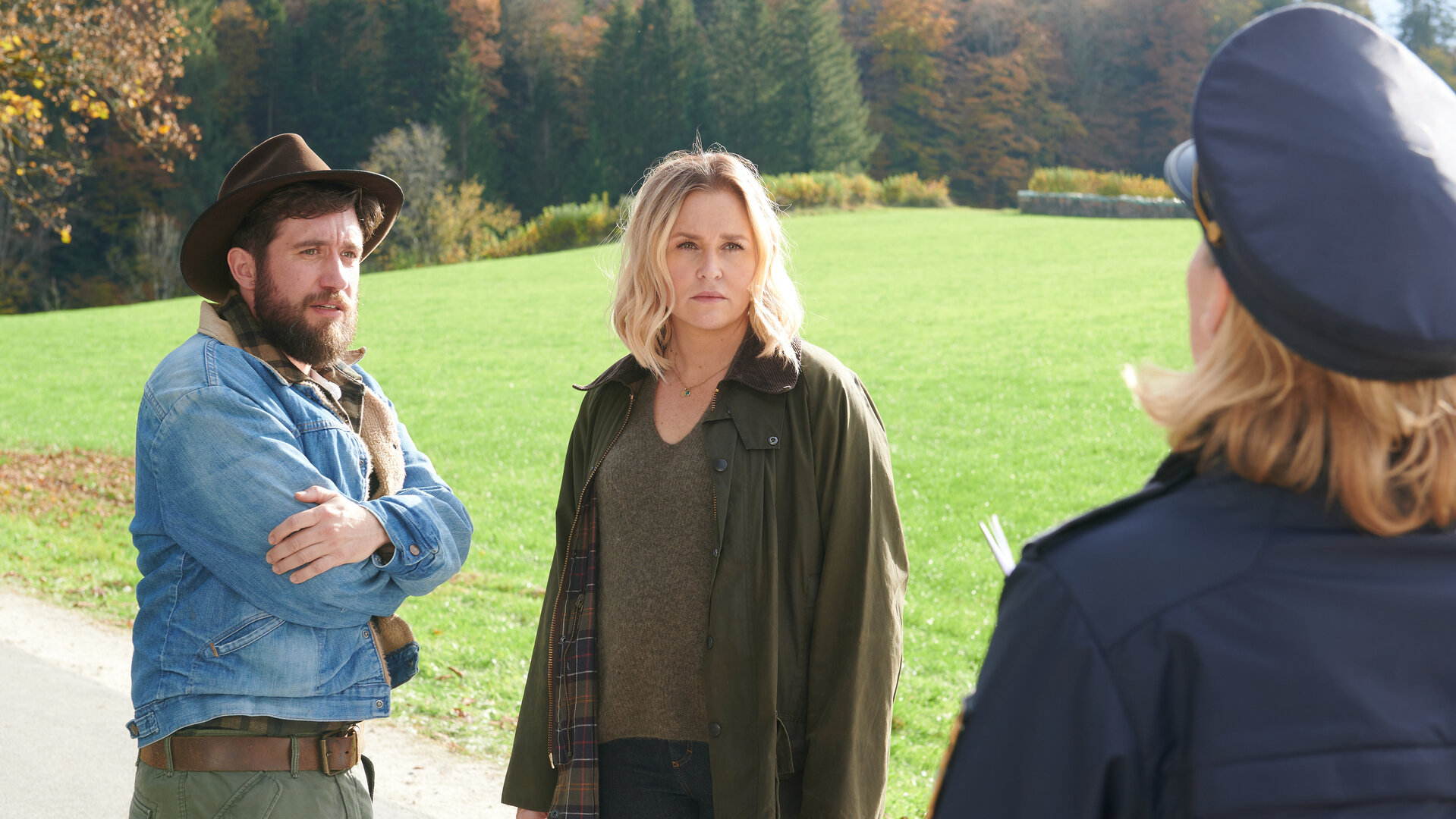 "Lena Lorenz - Gegen den Schmerz": Auf einer Straße stehen Schorschi Striebel (Sebastian Edtbauer) und Lena Lorenz (Judith Hoersch) und reden mit der Polizeibeamtin Anna Prückl (Sarah-Lavinia Schmidbauer), die von hinten zu sehen ist. Schorschi hat die Arme um seinen Oberkörper geschlungen und wirkt schockiert. Lena schaut die Polizistin ernst an.