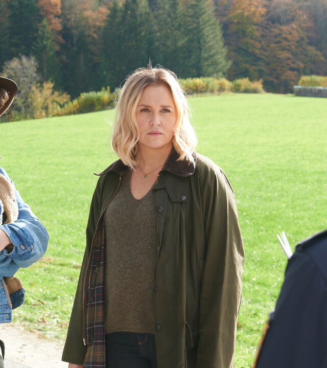 "Lena Lorenz - Gegen den Schmerz": Auf einer Straße stehen Schorschi Striebel (Sebastian Edtbauer) und Lena Lorenz (Judith Hoersch) und reden mit der Polizeibeamtin Anna Prückl (Sarah-Lavinia Schmidbauer), die von hinten zu sehen ist. Schorschi hat die Arme um seinen Oberkörper geschlungen und wirkt schockiert. Lena schaut die Polizistin ernst an.