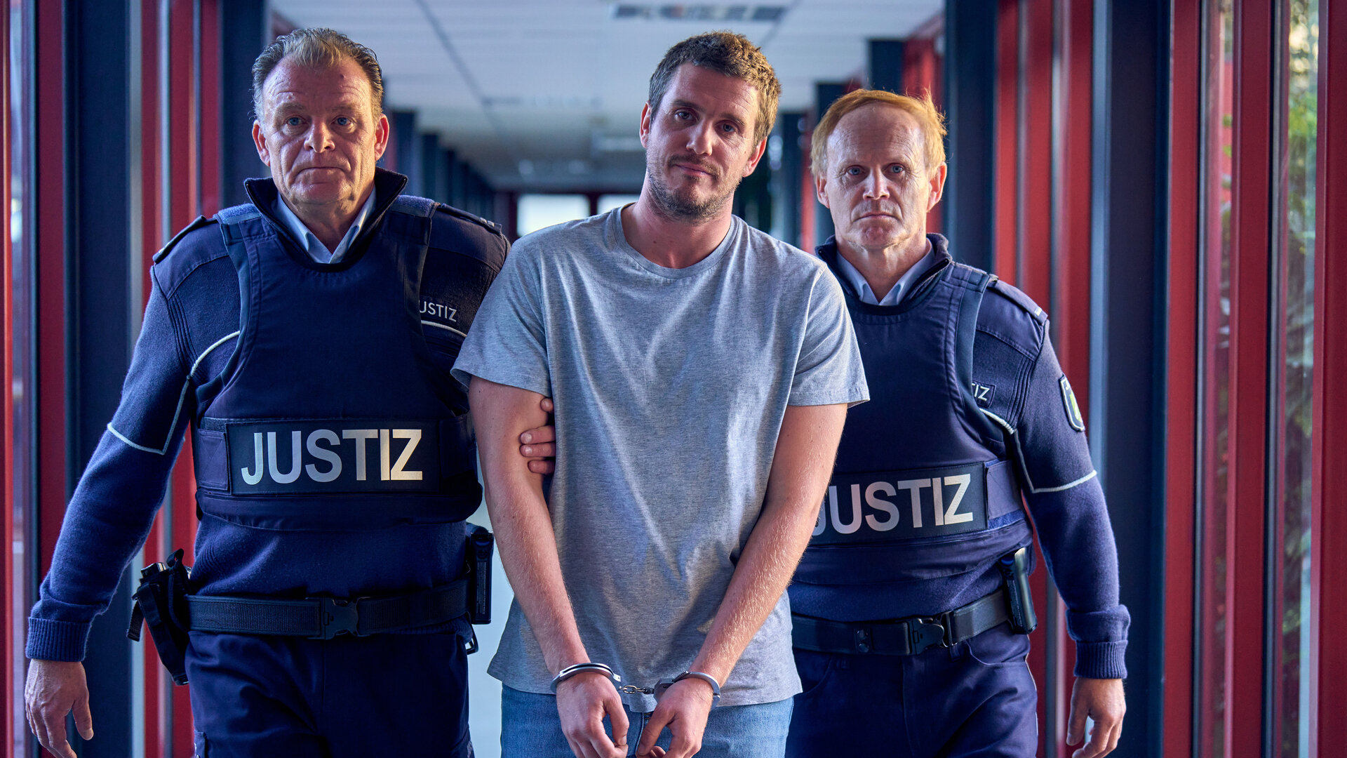 "SOKO Köln - Das Ultimatum": Alex Rohm (Max Bretschneider) wird in Handschellen von zwei uniformieren Polizisten (Komparsen) durch den Flur im Kommissariat geführt. Die Polizisten sehen ernst nach vorne, Alex Rohms Blick ist resigniert. Die drei Männer stehen frontal vor der Kamera.