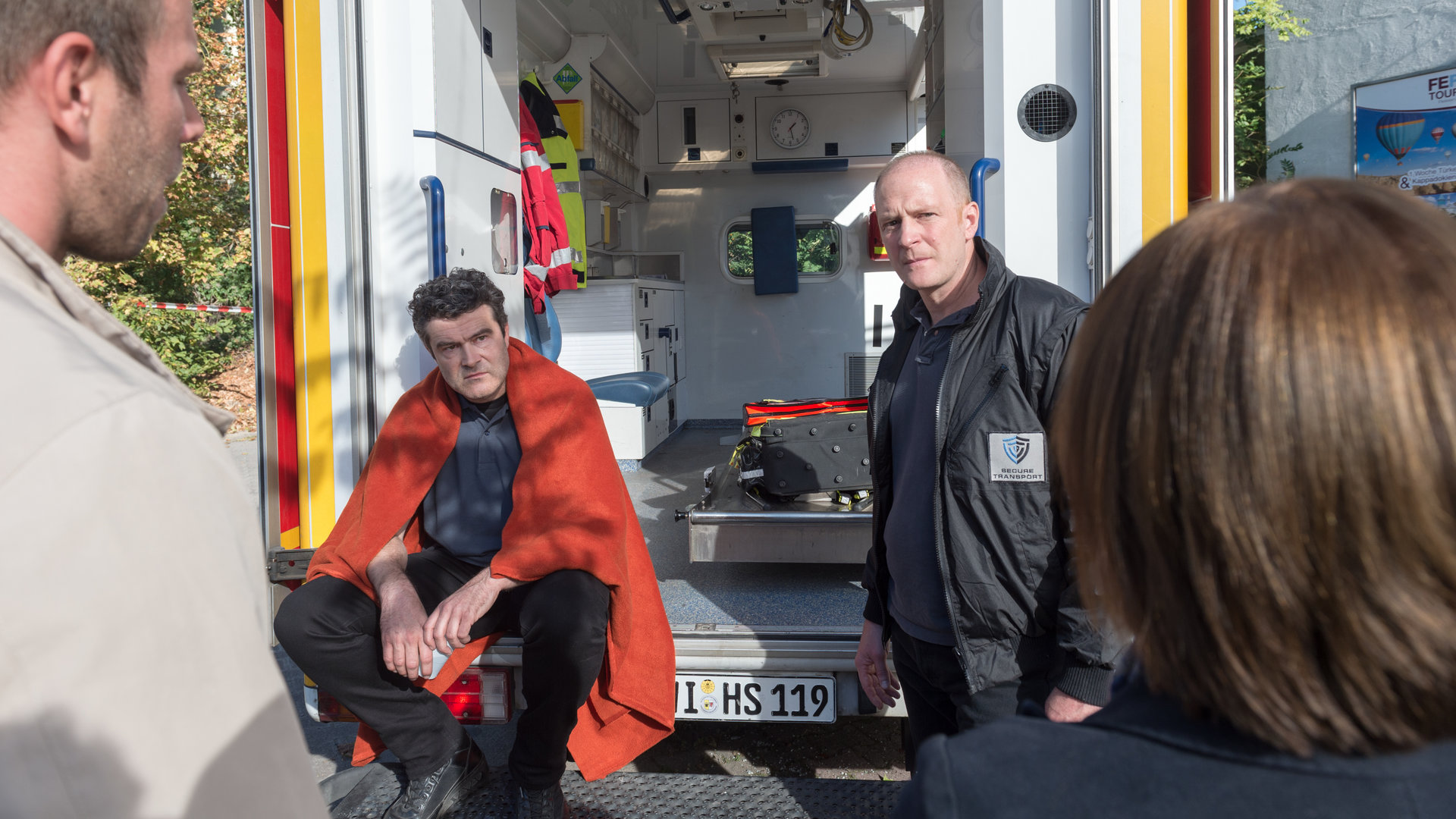 "SOKO Wismar – Zu spät": Lars Pöhlmann (Dominic Boeer) und Katrin Börensen (Claudia Schmutzler), nur von hinten zu sehen,  befragen die beiden Geldboten Jürgen Friedrich (Ecco Mylla) und Sven Keller (Christoph Jacobi). Keller sitzt, eine Decke um die Schultern, im geöffneten Krankenwagen, sein Kollege steht neben ihm. Beide schauen Katrin Börensen an.