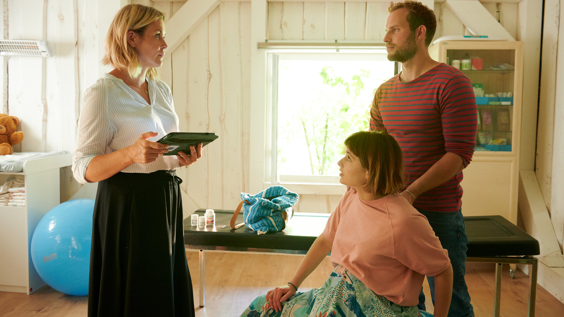 "Lena Lorenz - Krank vor Sorge": In Lena Lorenz' Praxis sitzt Tati Meisinger (Anouk Elias) auf einem großen Gymnastikball. Kaspar Hahn (Arne Löber) steht hinter ihr und stützt sie mit den Händen. Beide schauen Lena (Judith Hoersch) an, die ihnen gegenüber steht und sie ernst anblickt.
