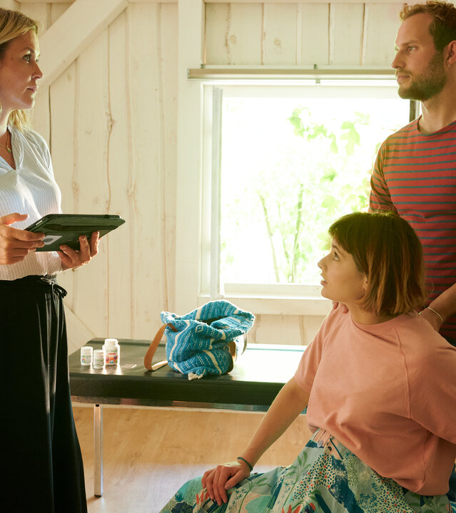 "Lena Lorenz - Krank vor Sorge": In Lena Lorenz' Praxis sitzt Tati Meisinger (Anouk Elias) auf einem großen Gymnastikball. Kaspar Hahn (Arne Löber) steht hinter ihr und stützt sie mit den Händen. Beide schauen Lena (Judith Hoersch) an, die ihnen gegenüber steht und sie ernst anblickt.