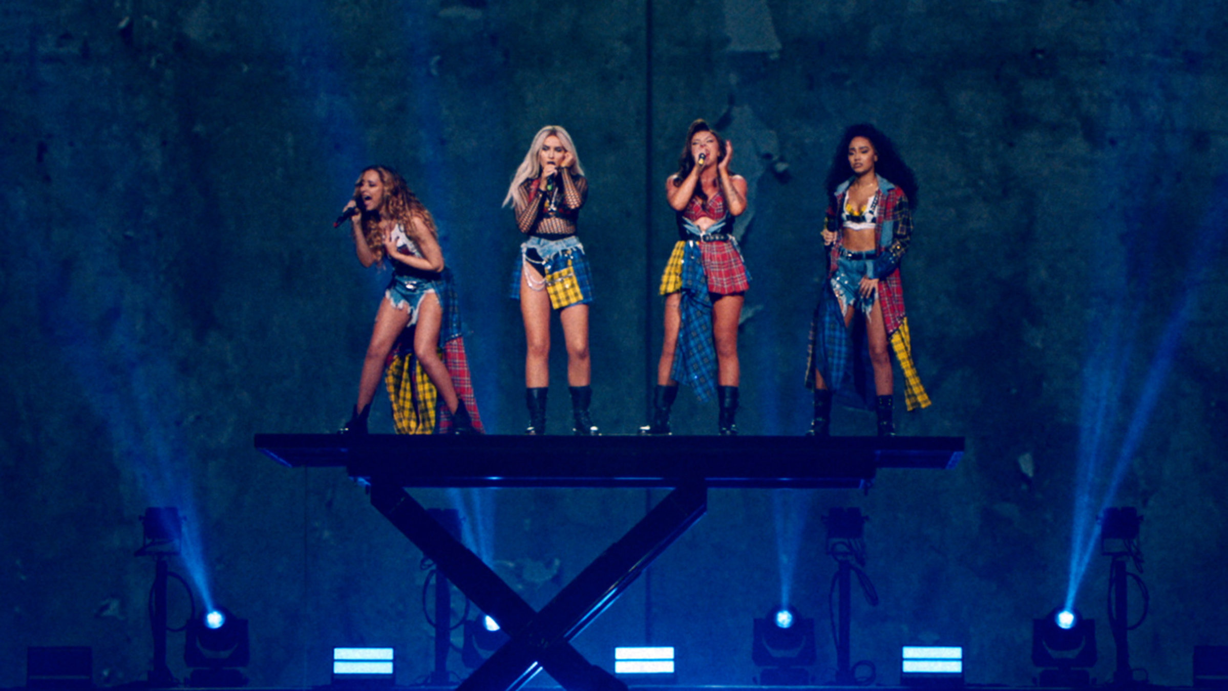 "Little Mix: LM 5 - The Tour Film": Vier Frauen stehen auf einer kleinen Hebe-Bühne und singen. Die Musikerinnen sind der Mittelpunkt des Bildes. Das Bühnenbild im Hintergrund ist blau. Scheinwerfer strahlen von unten nach oben. An der Decke, direkt über den vier Sängerinnen, ist eine dreieckige Lichtinstallation mit leuchtenden Glühbirnen zu sehen.