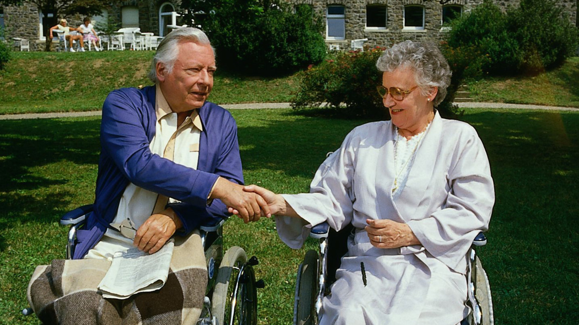 "Die Schwarzwaldklinik - Spätes Glück": Max Seidelbach (Hans Caninenberg) und Olga Herbold (Edda Seipel) sitzen in ihren Rollstühlen im Garten der Klinik und reichen sich lächelnd die Hand.
