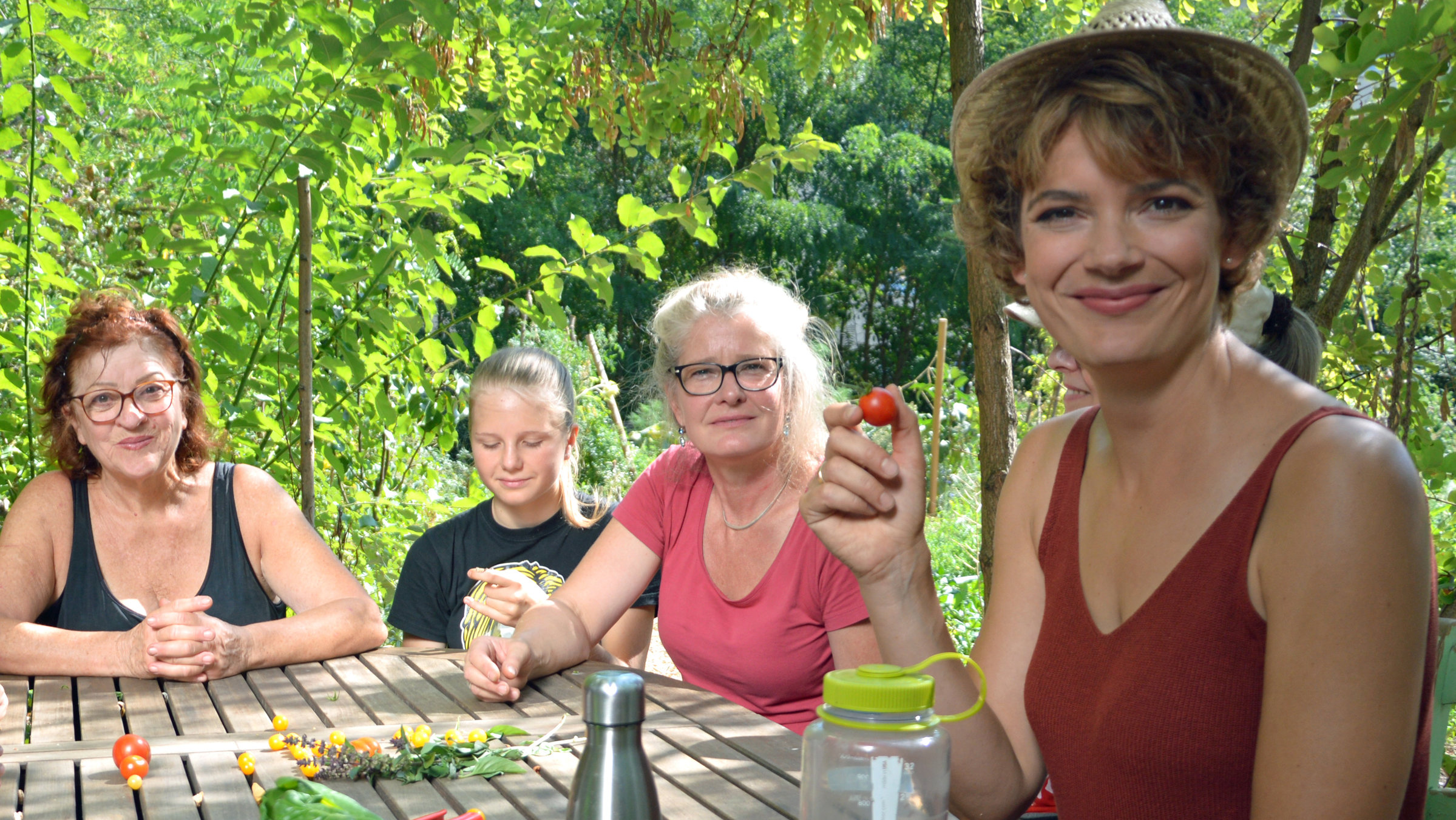 "Terra Xpress: Die Gemüseretter - Revolution in der Stadt": Moderatorin Lena Ganschow sitzt mit zwei Frauen und einem Mädchen an einem Holztisch. Sie lächelt in die Kamera und hält eine kleine Tomate zwischen Daumen und Zeigefinger. Auf dem Tisch liegen Blumen, Blätter und Tomaten. Außerdem steht eine Flasche mit Wasser und eine silberne Thermoskanne auf dem Tisch.