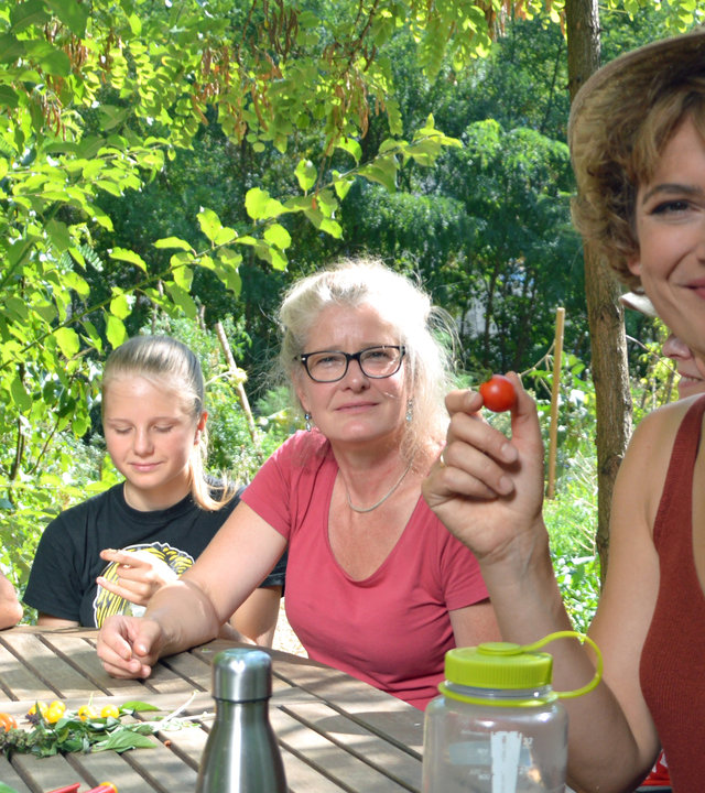 "Terra Xpress: Die Gemüseretter - Revolution in der Stadt": Moderatorin Lena Ganschow sitzt mit zwei Frauen und einem Mädchen an einem Holztisch. Sie lächelt in die Kamera und hält eine kleine Tomate zwischen Daumen und Zeigefinger. Auf dem Tisch liegen Blumen, Blätter und Tomaten. Außerdem steht eine Flasche mit Wasser und eine silberne Thermoskanne auf dem Tisch.