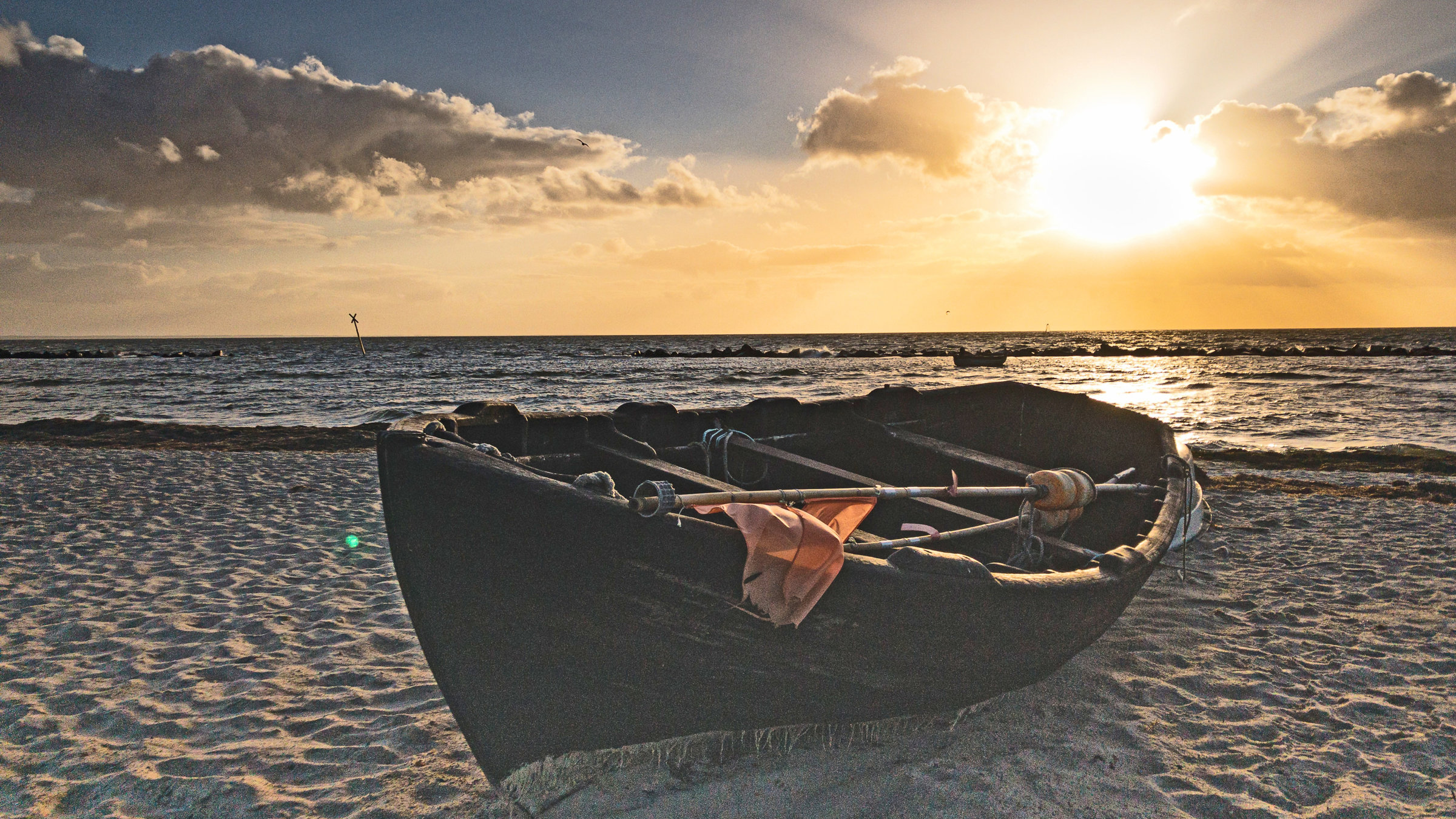 "Aussterbende Art": Abendstimmung am Strand. Ein hölzernes Fischerboot liegt im Sand. Im Hintergrund das Meer und die untergehende Sonne.