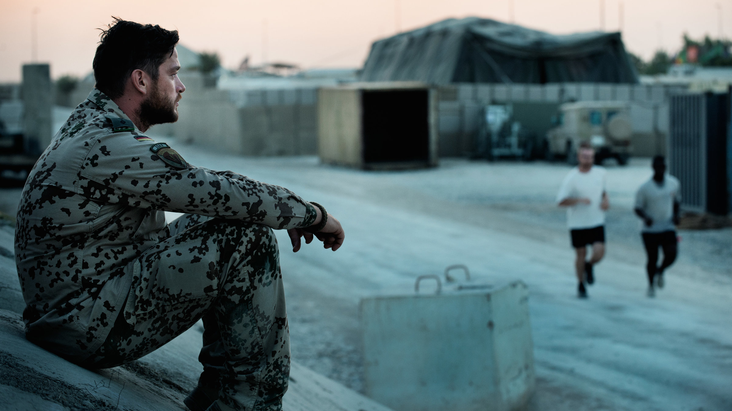 "Zwischen Welten": Jesper (Ronald Zehrfeld) sitzt in Bundeswehrkleidung auf einem Hügel vor einem Gelände, auf dem ein großes Zelt steht. Er blickt nachdenklich geradeaus. Zwei Jogger laufen an ihm vorbei.