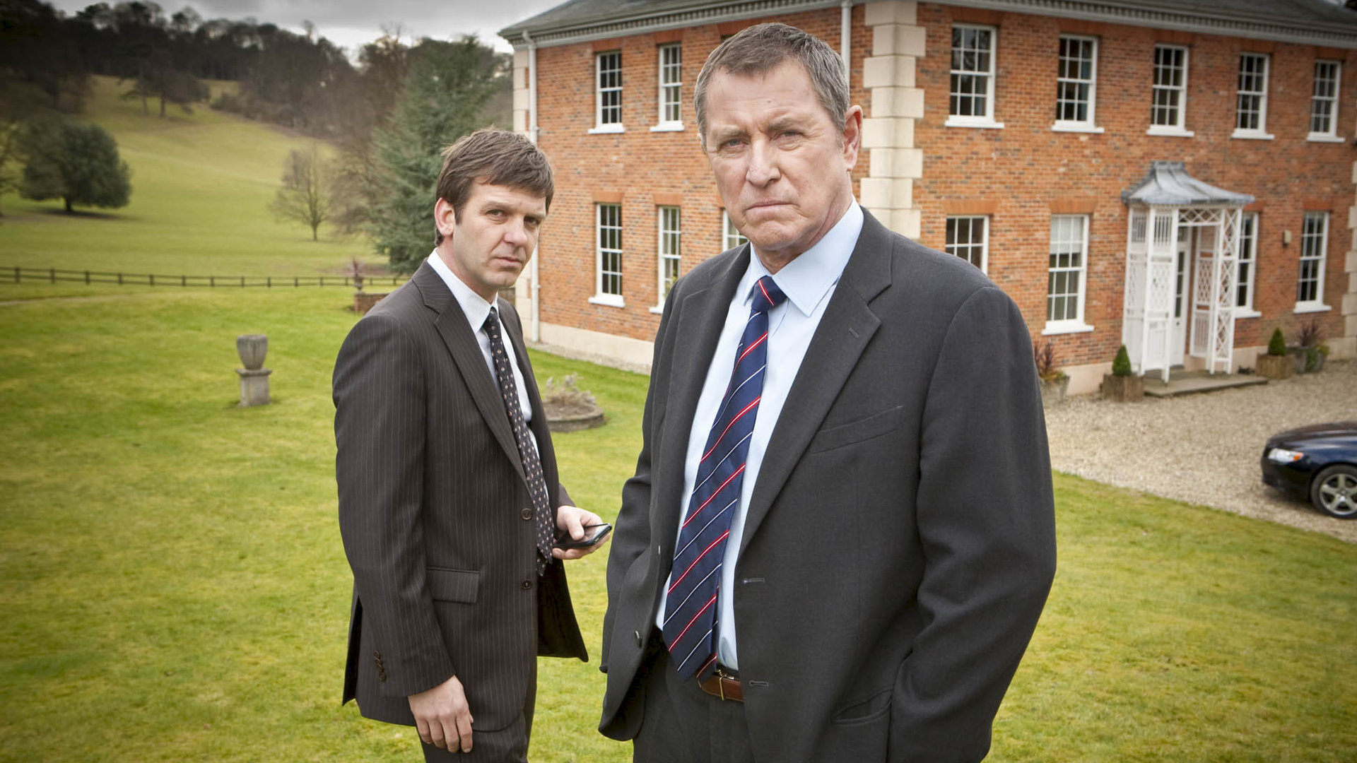"Inspector Barnaby - Über den Dächern von Chattham": DS Ben Jones (Jason Hughes) und DCI Tom Barnaby (John Nettles) stehen nebeneinander auf einer Wiese, hinter ihnen ist ein großes Backsteinhaus zu sehen.