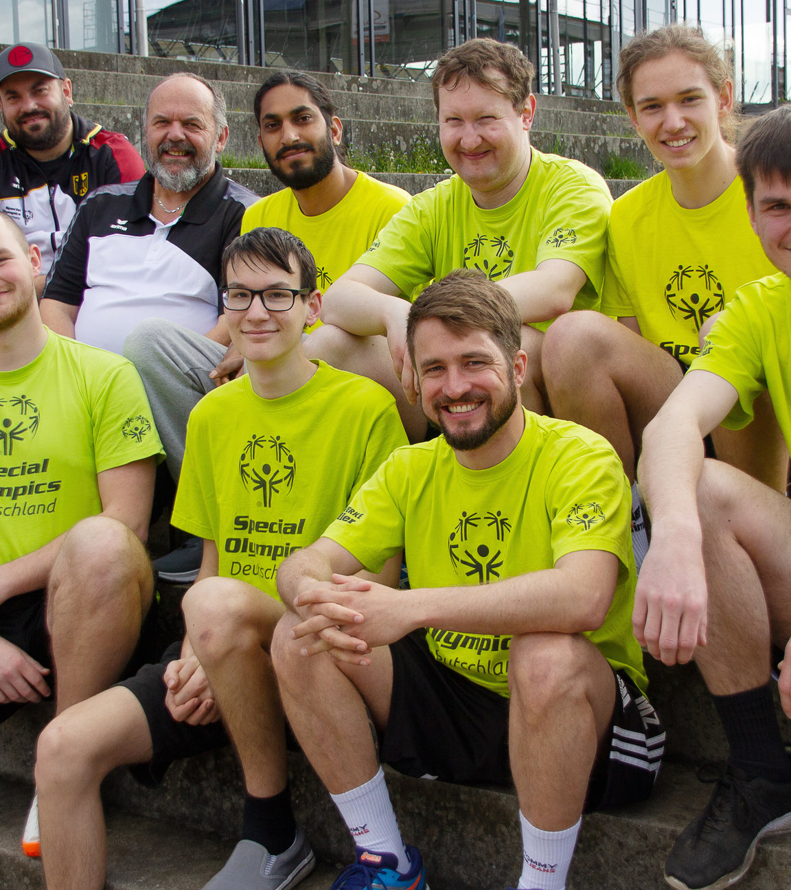 "einfach Mensch: Folge: Unified Volleyball – Im Team zu den Special Olympics 2023": Die Athleten, Unified-Partner und Trainer sitzen vor einer Volleyballhalle. Reihe unten von links: Donato Iasi, Maximo Winter, Sebastian Manz, Benedict Eninger, Dennis Kutzner; Reihe oben von links: Raphael Stäbler, Michael Stäbler, Timo Natzenberg, Matthias Aigner, Hannes Caspari