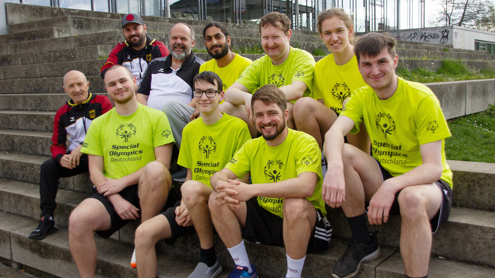 "einfach Mensch: Folge: Unified Volleyball – Im Team zu den Special Olympics 2023": Die Athleten, Unified-Partner und Trainer sitzen vor einer Volleyballhalle. Reihe unten von links: Donato Iasi, Maximo Winter, Sebastian Manz, Benedict Eninger, Dennis Kutzner; Reihe oben von links: Raphael Stäbler, Michael Stäbler, Timo Natzenberg, Matthias Aigner, Hannes Caspari