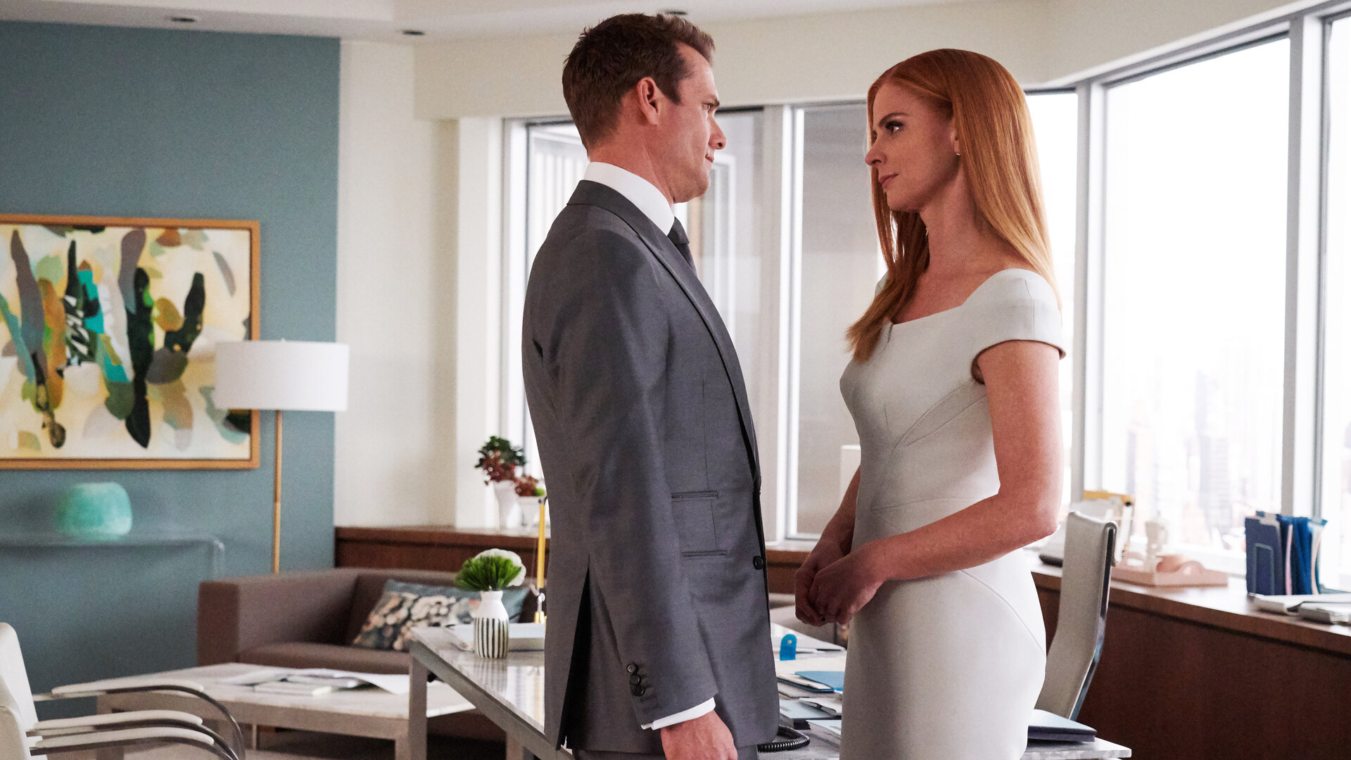"Suits - Windmühlen": Im Büro: Donna (Sarah Rafferty) und Harvey (Gabriel Macht) stehen sich sehr nah gegenüber und schauen sich dabei tief in die Augen. Im Hintergrund steht eine Sitzecke vor einer petrolfarbenen Wand, an der ein modernes abstraktes Gemälde hängt.