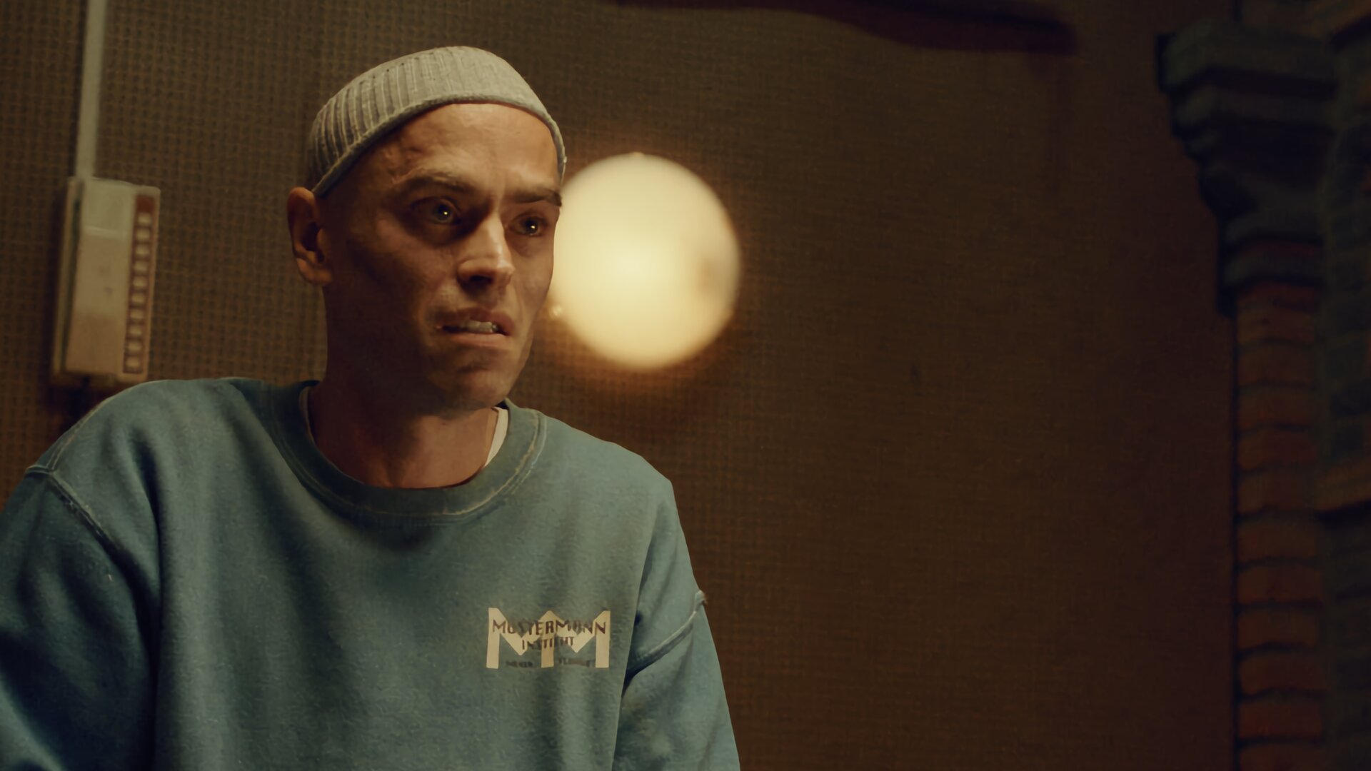 "Moresnet - The Book of Death: Der zweite Upload": Daan Schotz (Arend Pinoy) sitzt in einem Raum an einem Tisch und unterhält sich mit einer Person, die ihm gegenübersitzt, aber nicht zu erkennen ist. Auf dem Tisch liegen Unterlagen. Daan wirkt verschlossen. Er trägt einen verschmutzten grünblauen Pulli und eine dünne Strickmütze.