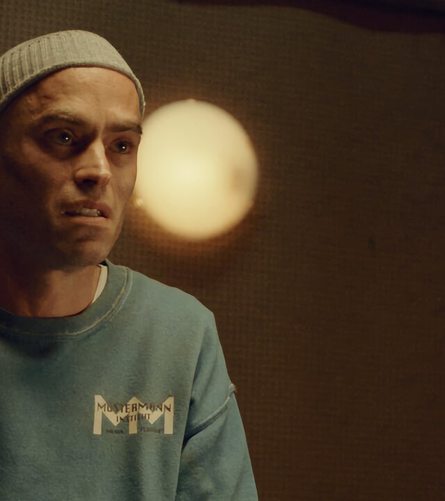 "Moresnet - The Book of Death: Der zweite Upload": Daan Schotz (Arend Pinoy) sitzt in einem Raum an einem Tisch und unterhält sich mit einer Person, die ihm gegenübersitzt, aber nicht zu erkennen ist. Auf dem Tisch liegen Unterlagen. Daan wirkt verschlossen. Er trägt einen verschmutzten grünblauen Pulli und eine dünne Strickmütze.