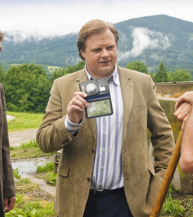 "Die Rosenheim-Cops - Späte Rache": Die Rosenheim-Cops Christian Lind (Tom Mikulla) und Tobias Hartl (Michael A. Grimm) befragen Bauer Wilfried Duch (Johann Schuler), der sich auf einen Holzstab stützt. Hartl zeigt Duch seine Polizeimarke. Im Hintergrund sind bewaldete Berge zu sehen.