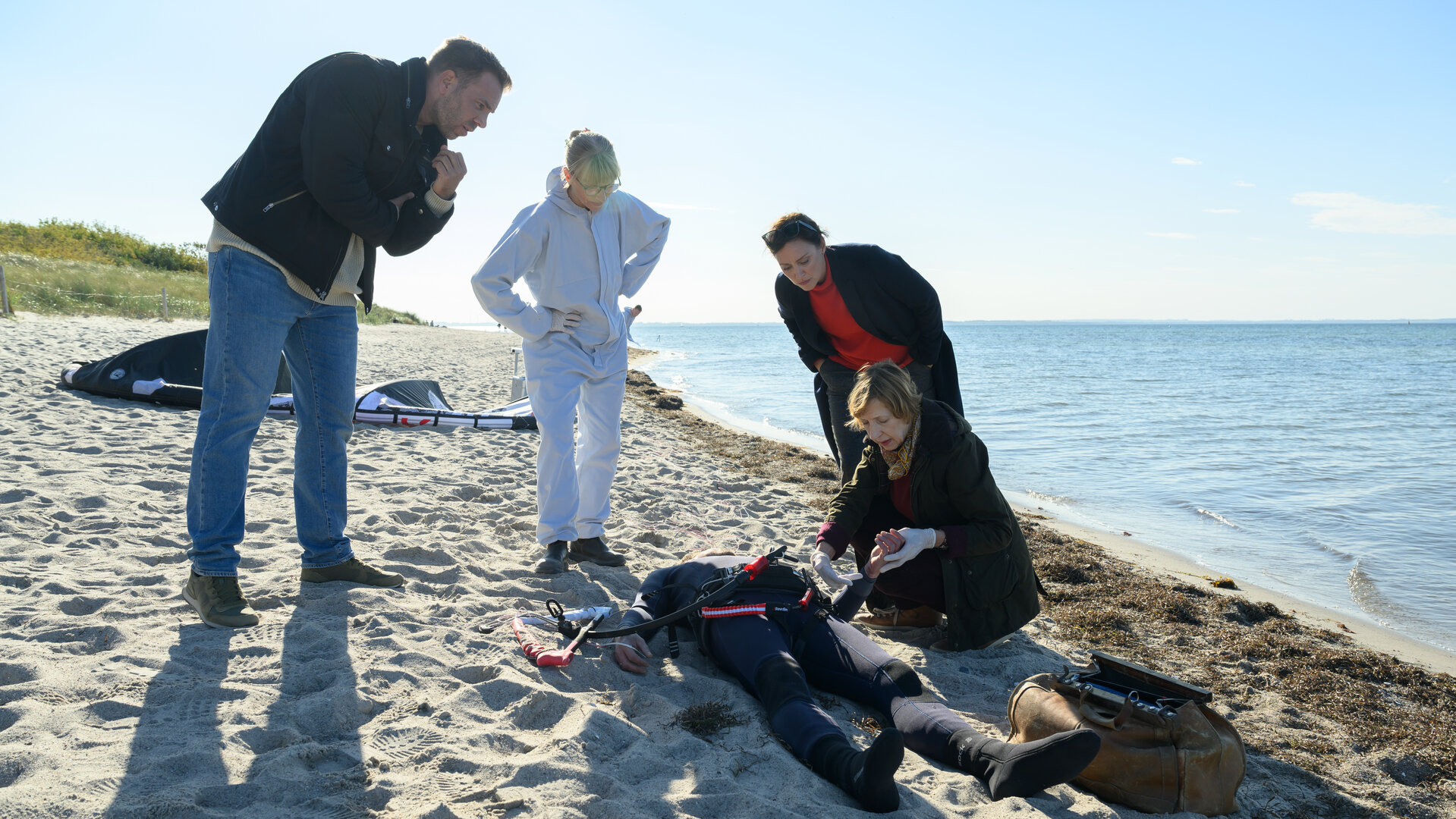 „SOKO Wismar – Kite in den Tod“: Die Leiche von Henning Waslund (Adrian Otzdorff) liegt nahe der Wasserkante am Strand. Lars Pöhlmann (Dominic Boeer), Roswitha Prinzler (Silke Matthias) und Karoline Joost (Nike Fuhrmann) stehen bei Dr. Helene Sturbeck (Katharina Blaschke), die den Leichnam untersucht. Das Kitesurf-Equipment liegt im Hintergrund im Sand.