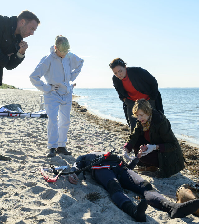 „SOKO Wismar – Kite in den Tod“: Die Leiche von Henning Waslund (Adrian Otzdorff) liegt nahe der Wasserkante am Strand. Lars Pöhlmann (Dominic Boeer), Roswitha Prinzler (Silke Matthias) und Karoline Joost (Nike Fuhrmann) stehen bei Dr. Helene Sturbeck (Katharina Blaschke), die den Leichnam untersucht. Das Kitesurf-Equipment liegt im Hintergrund im Sand.