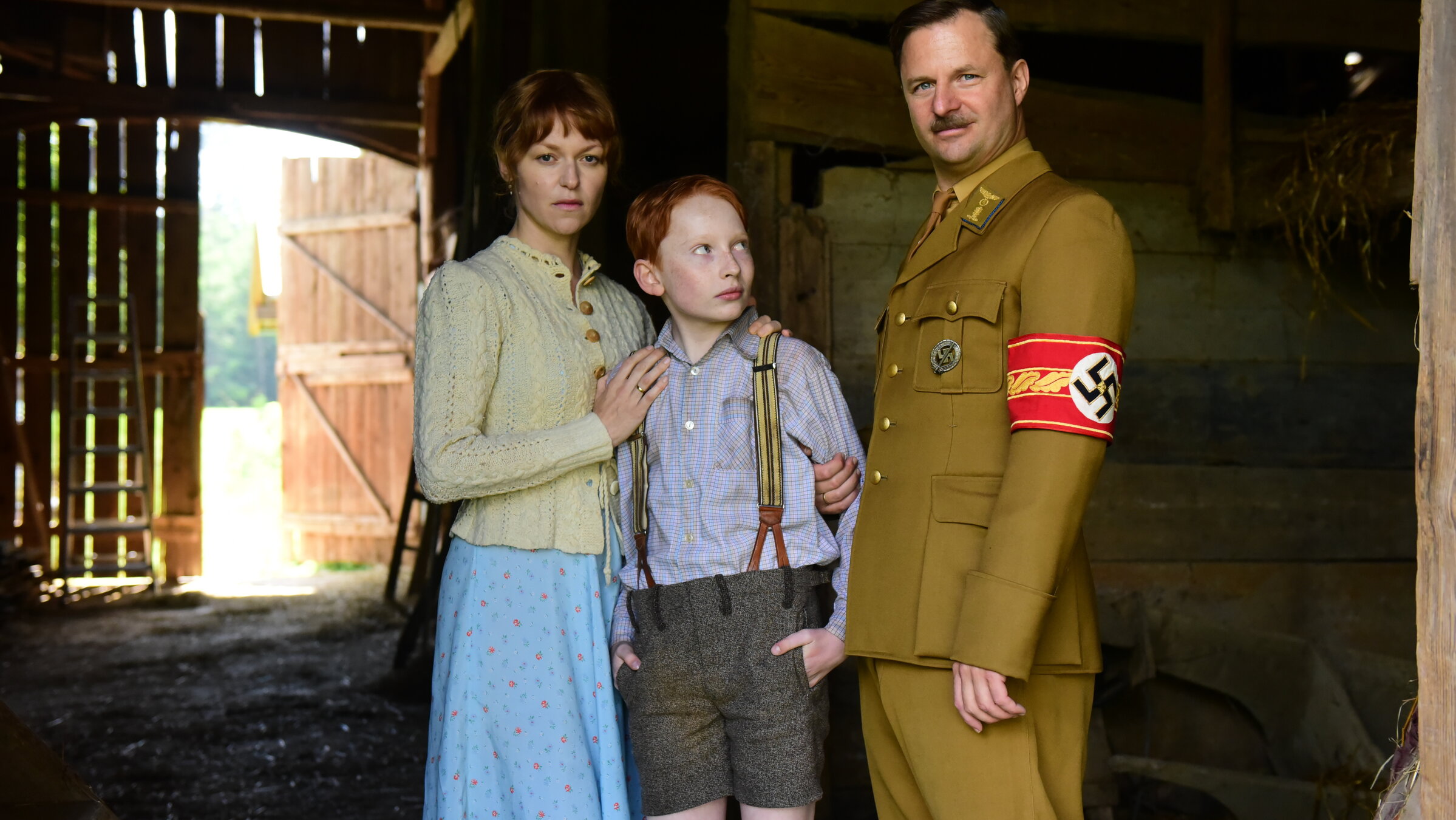 "Das Glaszimmer": In einem Stall stehen eine Frau (Lisa Wagner), die mit ernstem Blick ihren Sohn (Xari Wimbauer) im Arm hält. Dieser schaut ängstlich zu einem Mann (Philipp Hochmair) auf, der ihn grinsend und überheblich am Arm festhält. Er trägt die Hakenkreuzbinde am Arm.