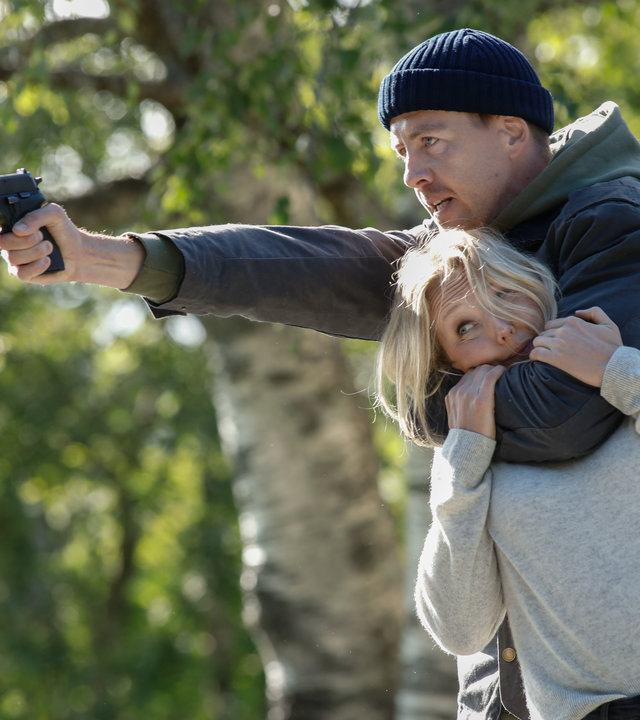 "Furia (1)": Im Wald hält Asgeir (Pål Sverre Hagen) mit seinem linken Arm Ragna (Ine Marie Wilmann) im Schwitzkasten, während er mit der Pistole in seiner rechten Hand auf jemanden außerhalb des Bildes zielt.