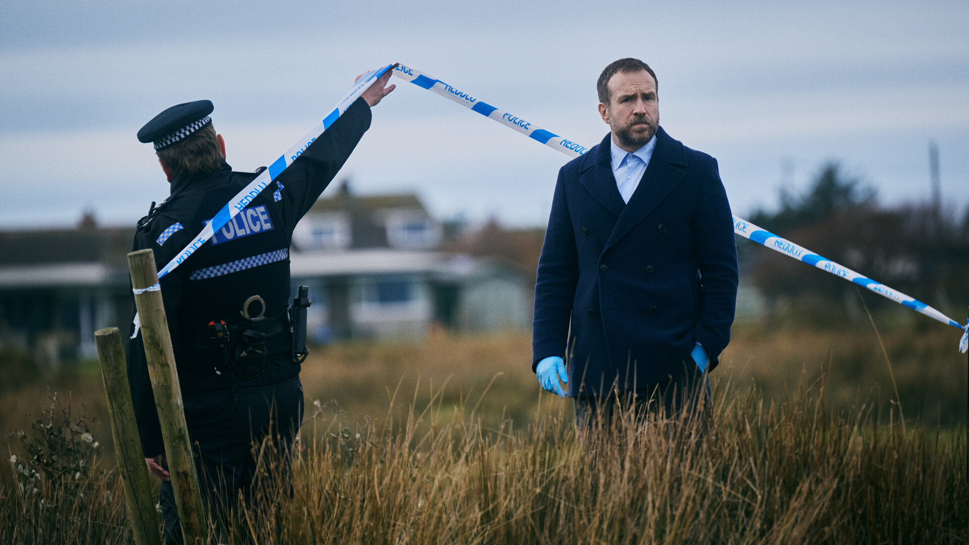 "Under Salt Marsh - Tod in den Salzwiesen - Ein grauenhafter Fund": Detective Eric Bull (Rafe Spall) steht im Freien auf einem Feld, das mit hohem Schlickgras bewachsen ist. Er trägt eine dunkle Jacke und Plastikhandschuhe. Neben ihm hält ein Polizist in Uniform (Komparse) ein Absperrband hoch.