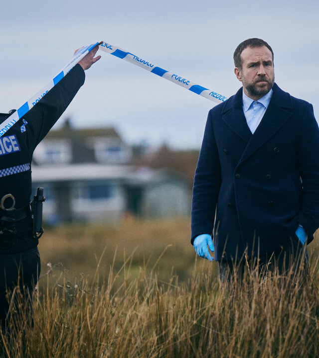 "Under Salt Marsh - Tod in den Salzwiesen - Ein grauenhafter Fund": Detective Eric Bull (Rafe Spall) steht im Freien auf einem Feld, das mit hohem Schlickgras bewachsen ist. Er trägt eine dunkle Jacke und Plastikhandschuhe. Neben ihm hält ein Polizist in Uniform (Komparse) ein Absperrband hoch.
