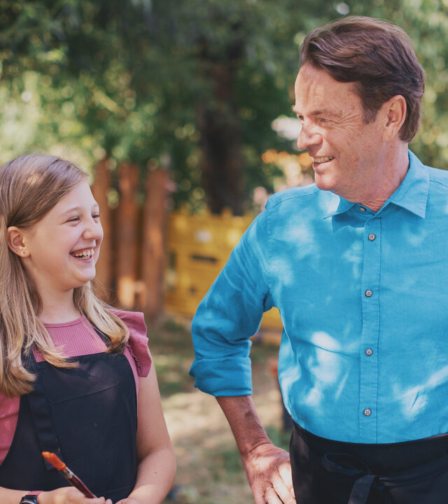 "Aktion Mensch - Glückszahlen der Woche": Rudi Cerne und ein Mädchen in dunkler Schürze mit einem Pinsel stehen nebeneinander in einem Gartenbereich. Sie schauen sich lachend an. Im Hintergrund stehen weitere Kinder zwischen bunten bemalten Leinwänden.